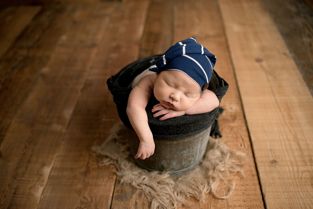 a baby lays posed in a bucket against hardwood planks during a newborn photography sessions