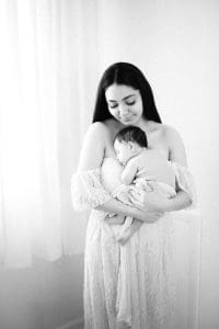 Mom stands in front of softly lit window holding daughter