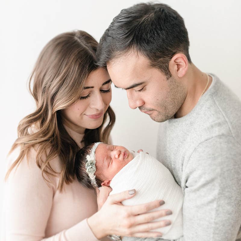 a mother and father stand huddled together holding their swaddled newborn baby