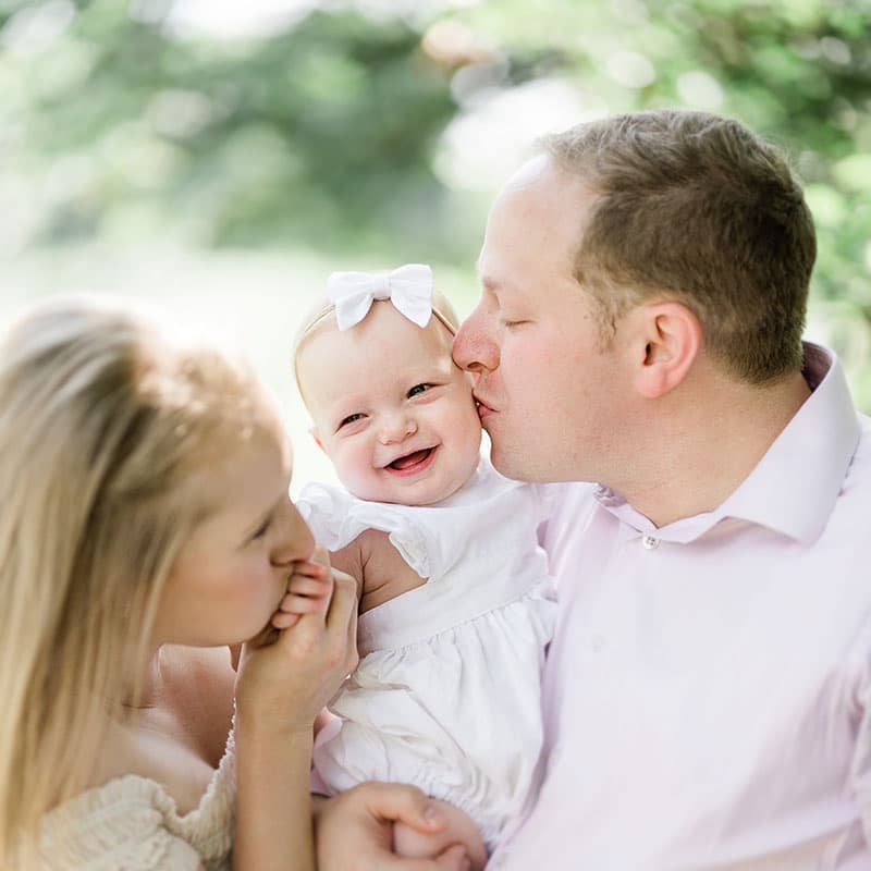 A baby smiles at the camera as dad hold her and mom kisses her little hand