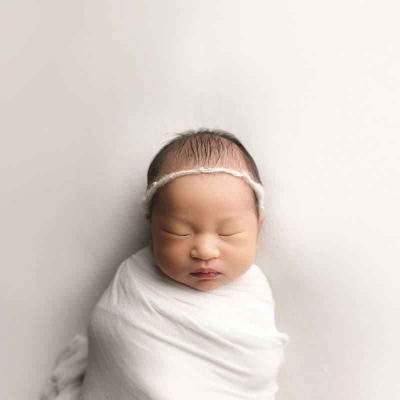 A newborn baby lays swaddled against a white seamless blanket