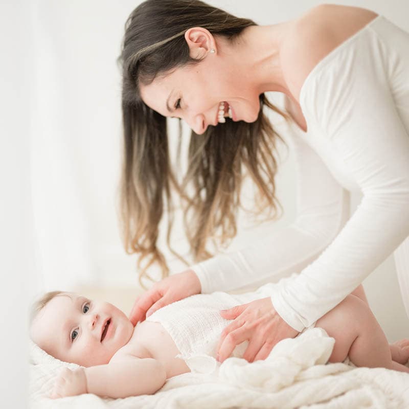 a baby lays on a blanket in front a large soft window while mother looks down smiling