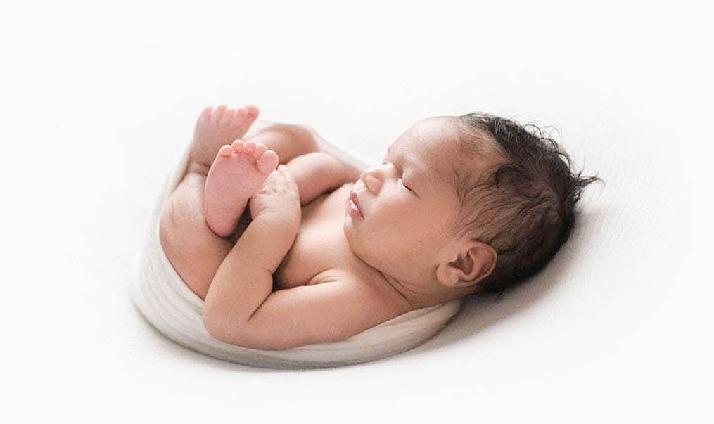 A newborn baby lays swaddled on a white blanket under soft white light